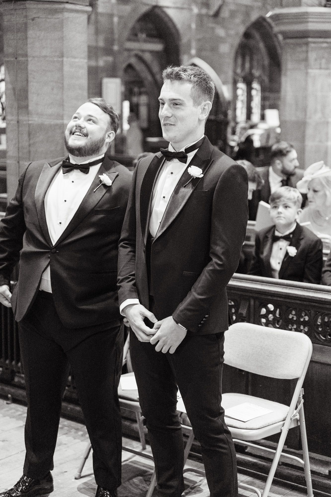 Two groomsmen in black tuxedos share a candid moment during a wedding ceremony in a historic church setting.
