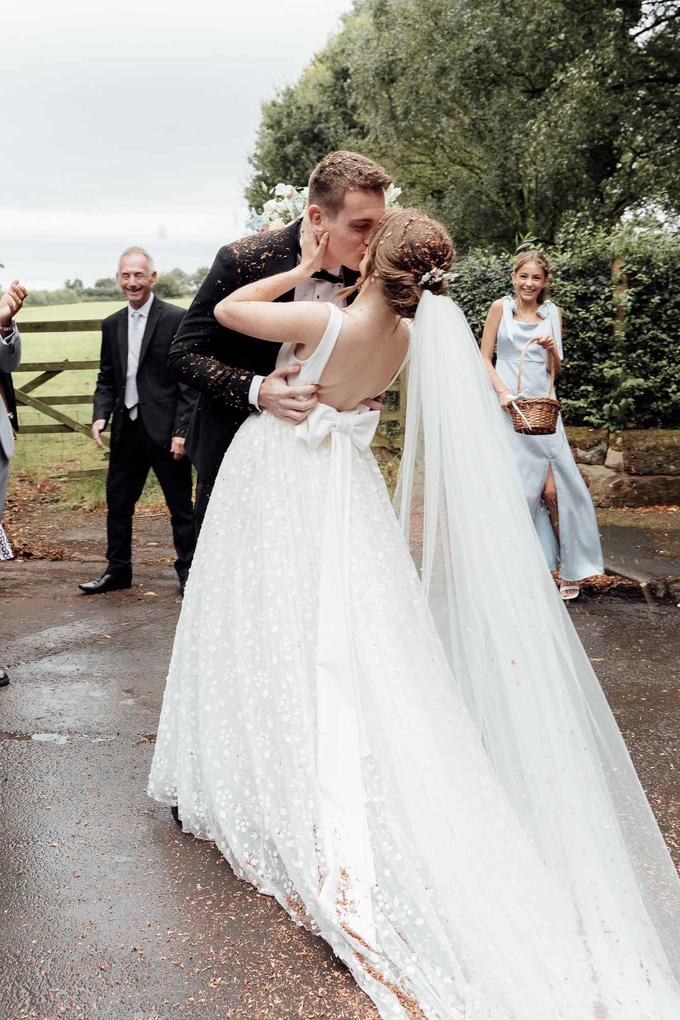 A wedding party walking along a gravel path surrounded by greenery and trees at an outdoor venue.