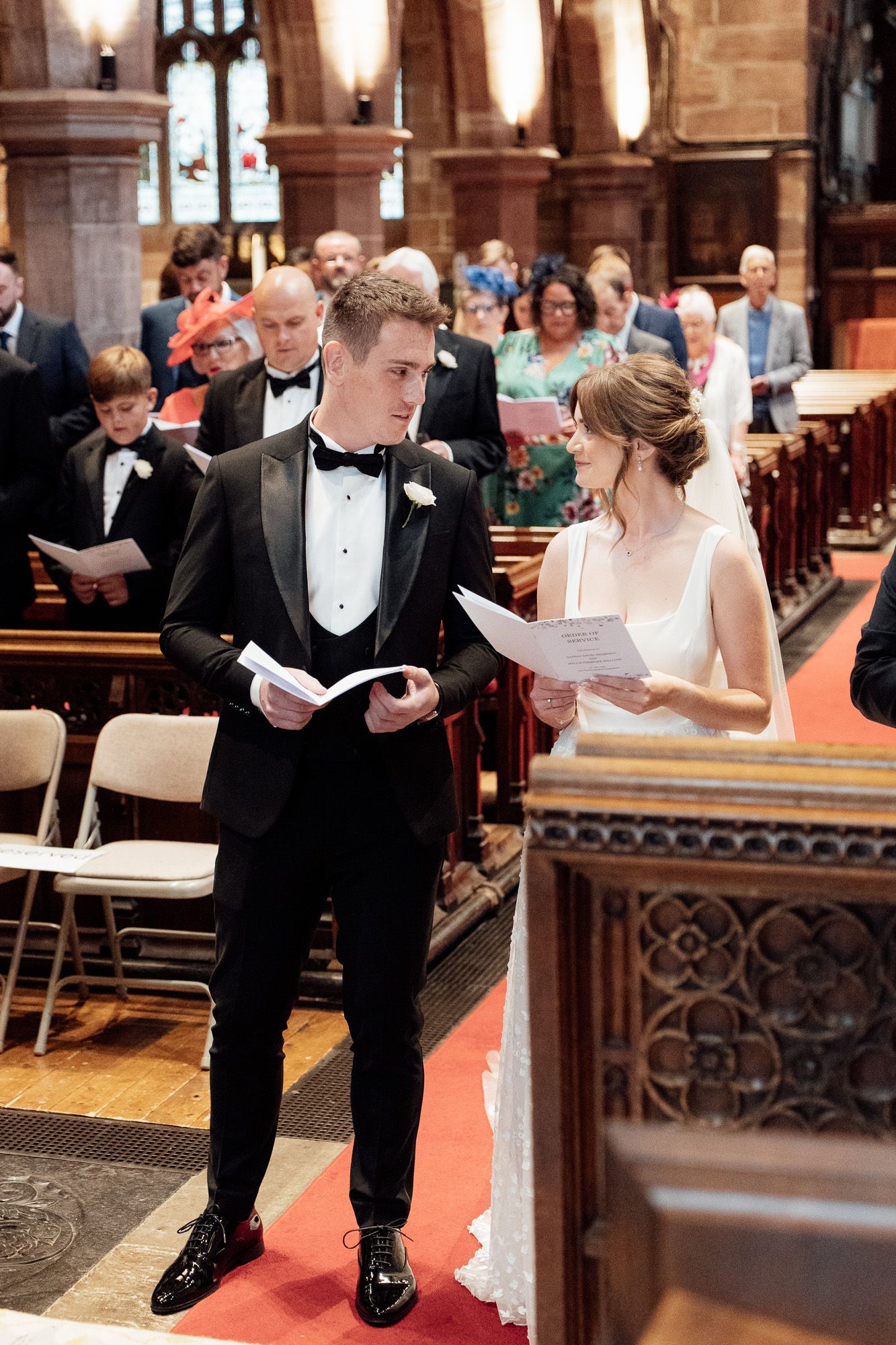 A couple in formal wedding attire reads from programs during a traditional church ceremony on a red carpet.