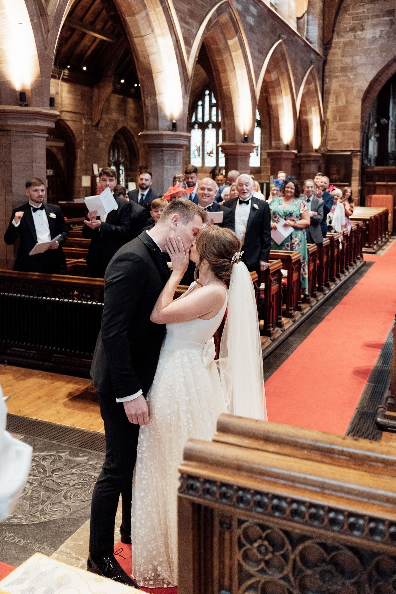 A couple shares their first kiss during a wedding ceremony inside a beautiful gothic church with arched ceilings and stained glass.