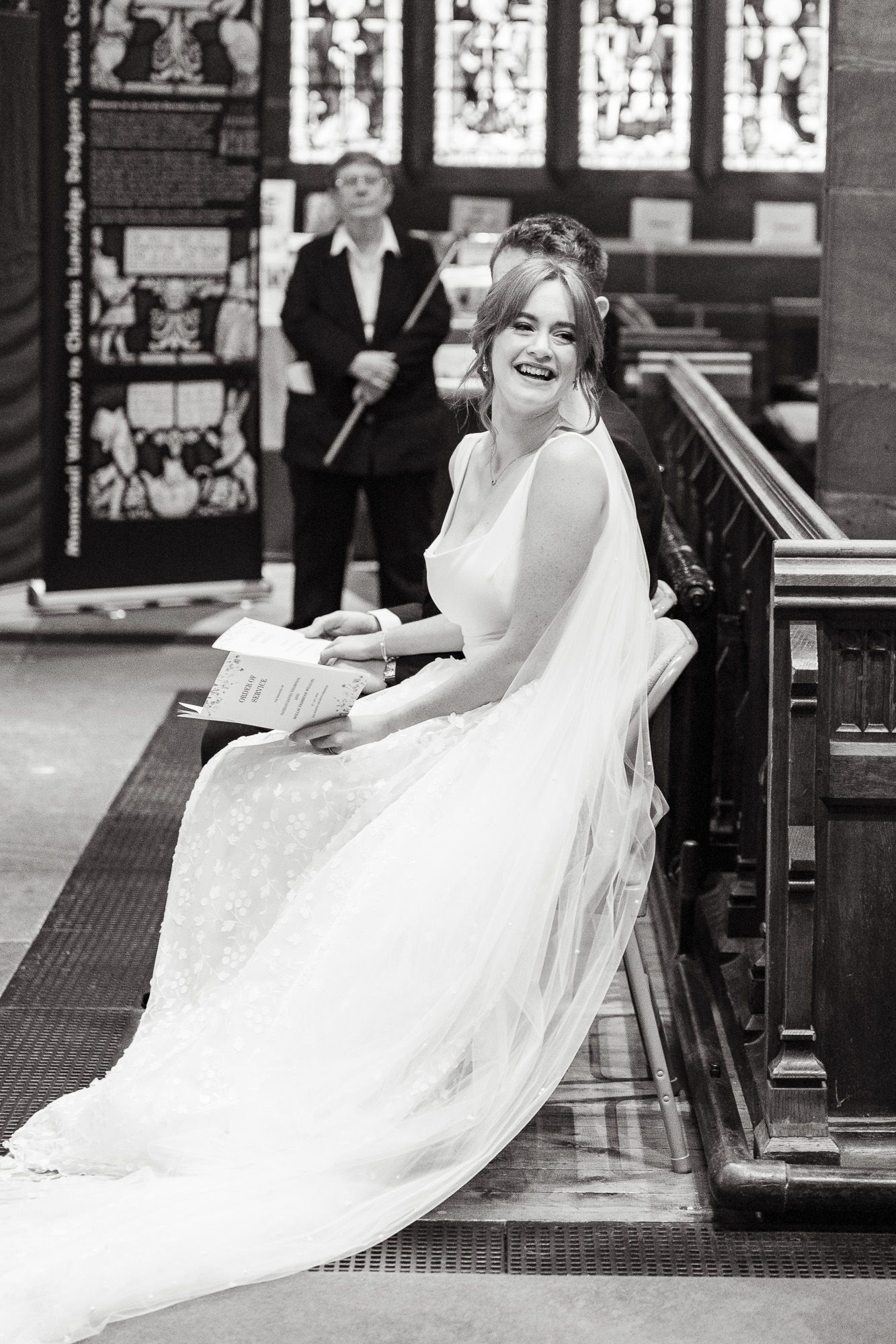 A joyful bride in a flowing white dress sits on church pews while laughing during a wedding ceremony.