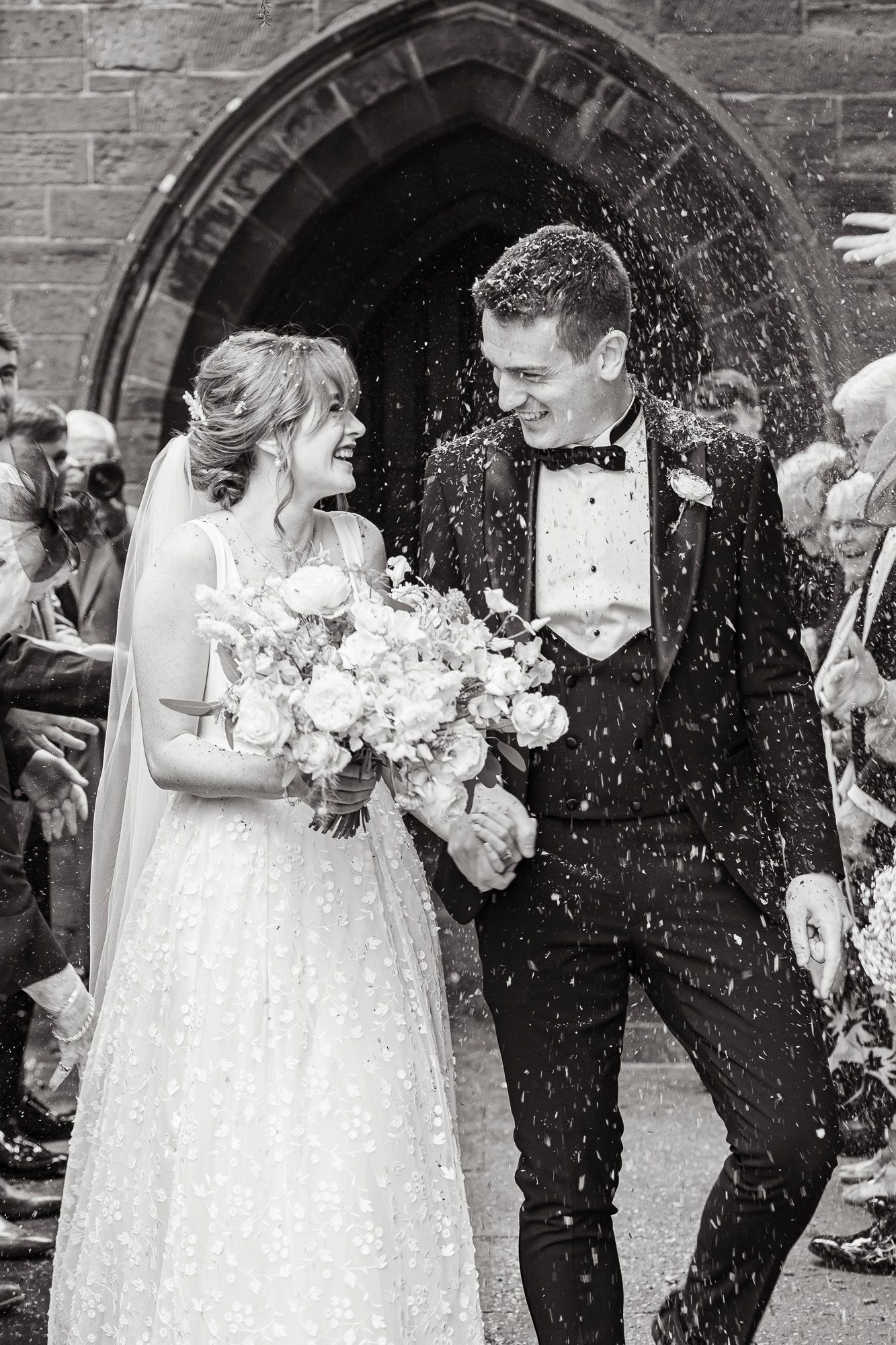A black and white wedding photo showing a couple joyfully exiting a church under a shower of confetti.