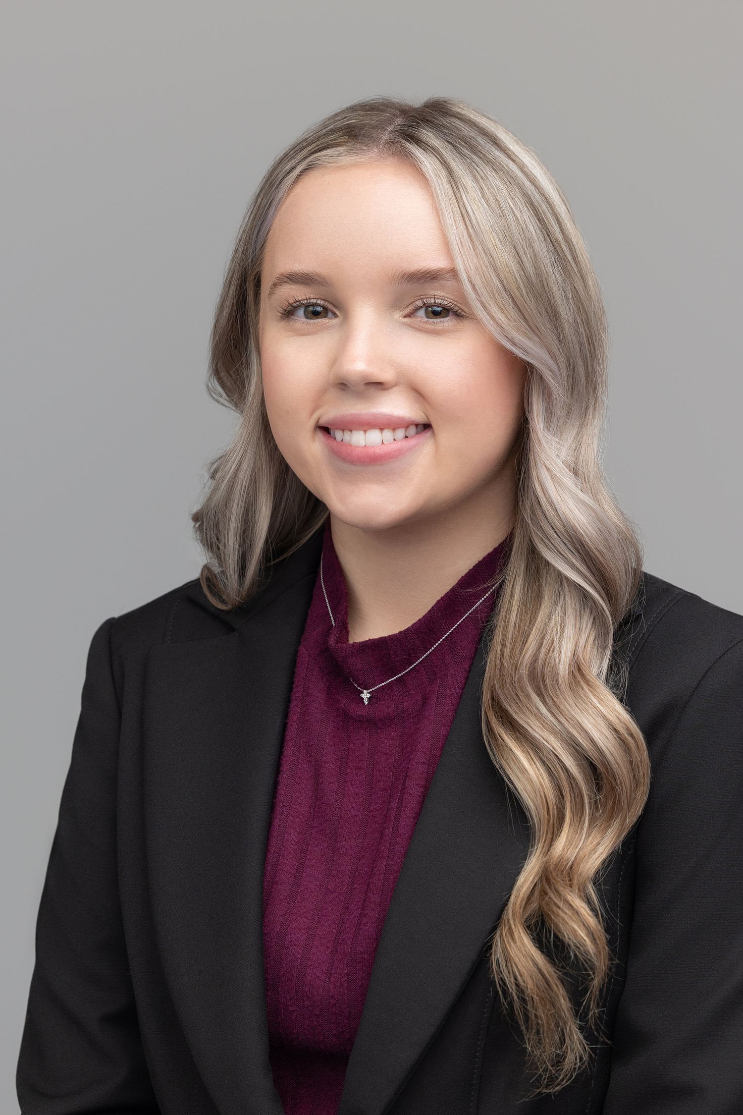 Professional headshot of a person with long wavy blonde hair wearing a black blazer and burgundy top against a grey background.