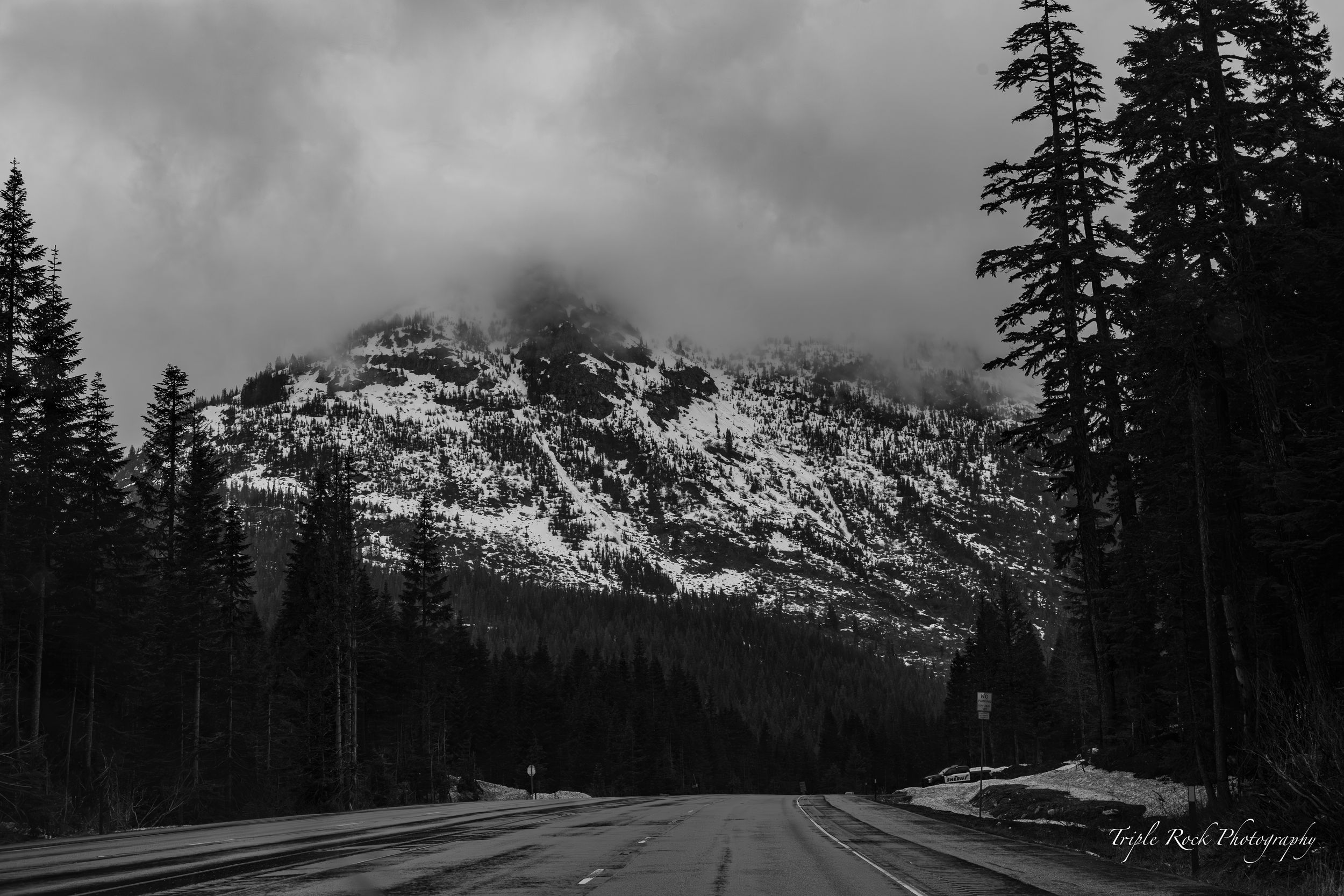 A snow-covered mountain peak looms over a winding forest road beneath dark storm clouds in black and white. Photographed by Jon Rock