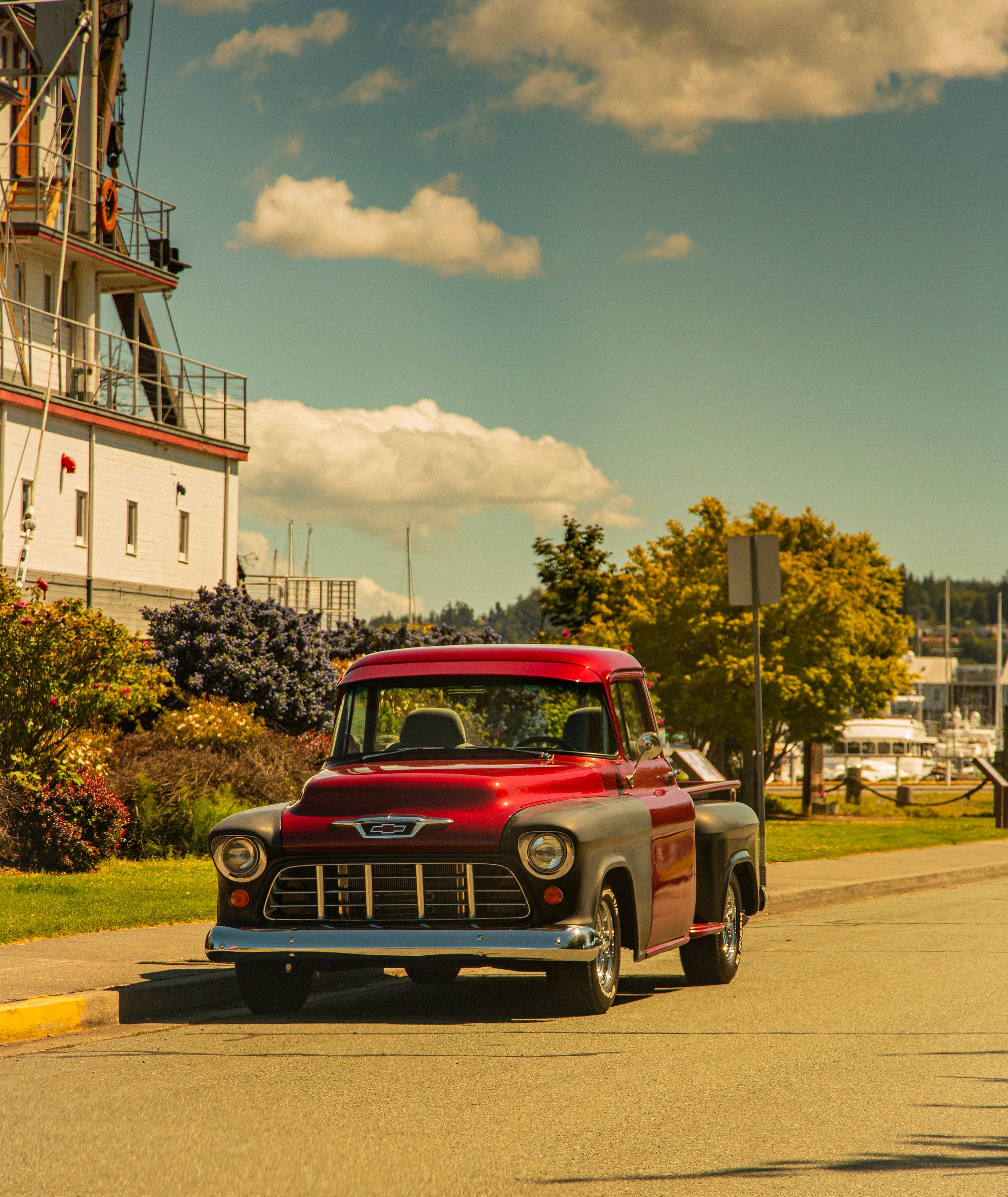 A vintage red 1950s Chevrolet pickup truck parked near a harbor marina with autumn trees and cloudy skies.