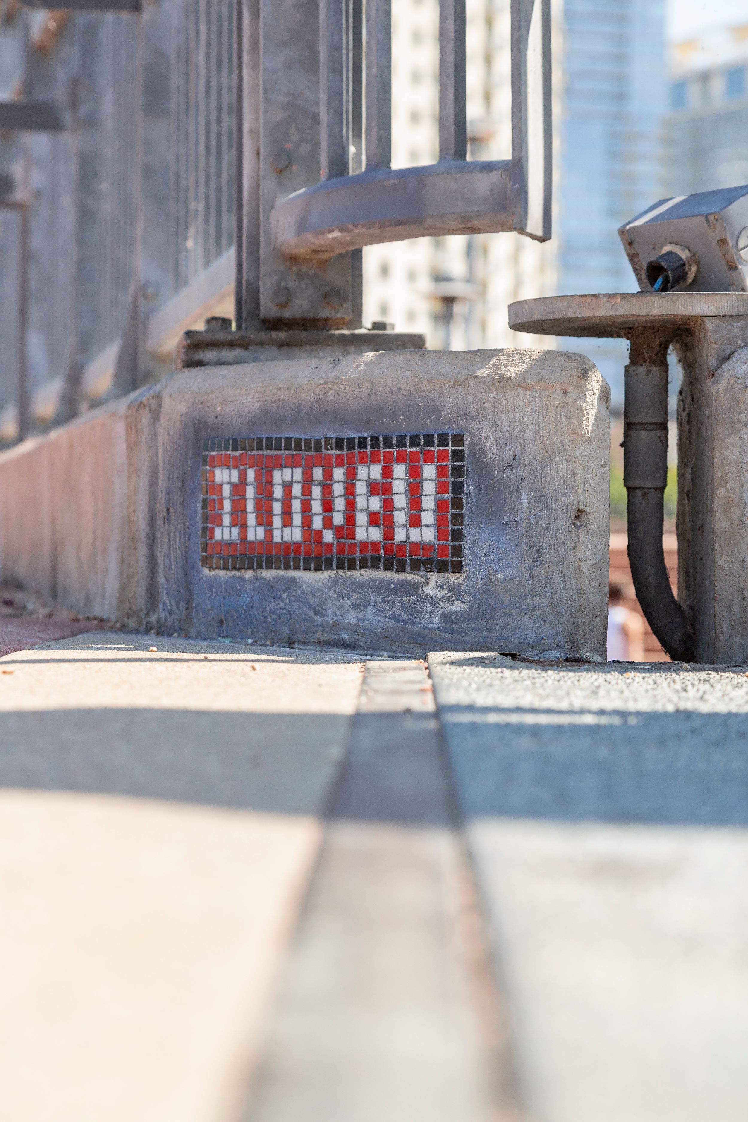 Close-up of a red mosaic 'LOVED' sign embedded in concrete with blurred city background.