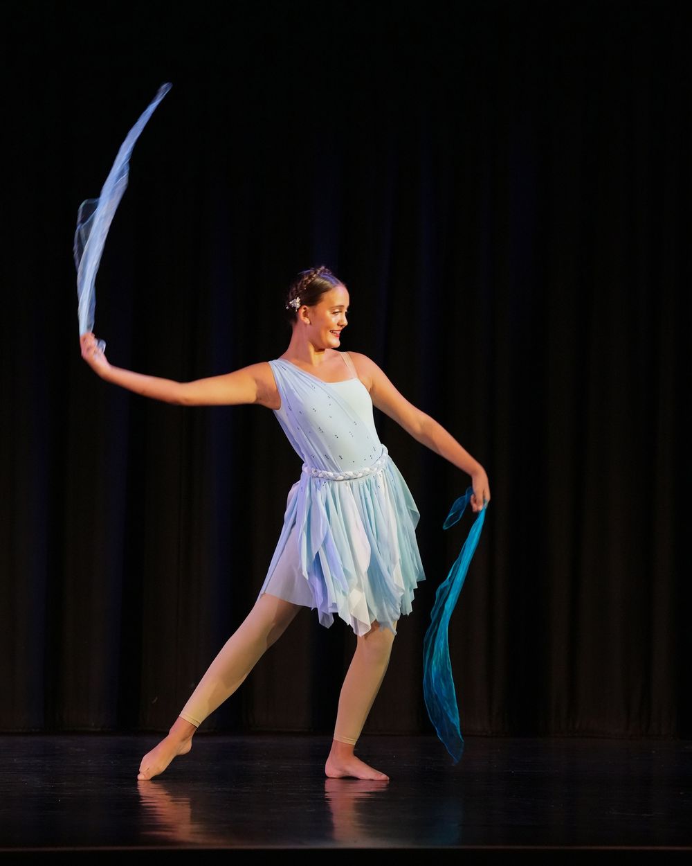 Young dancer in light blue costume gracefully performs on stage, flowing fabric ribbons in motion under spotlights.