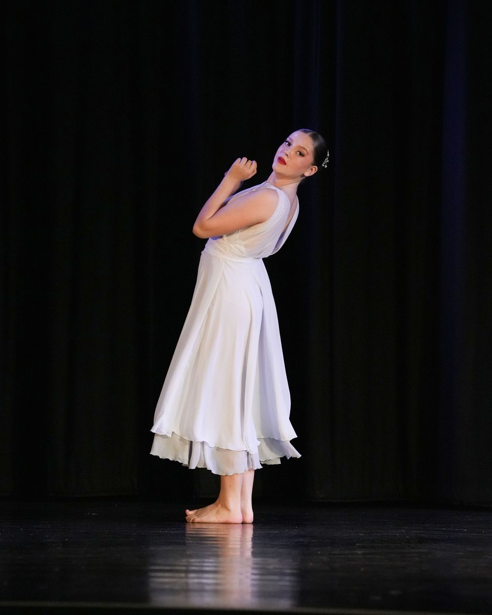 Young dancer in white dress performs graceful contemporary ballet on stage under spotlight.