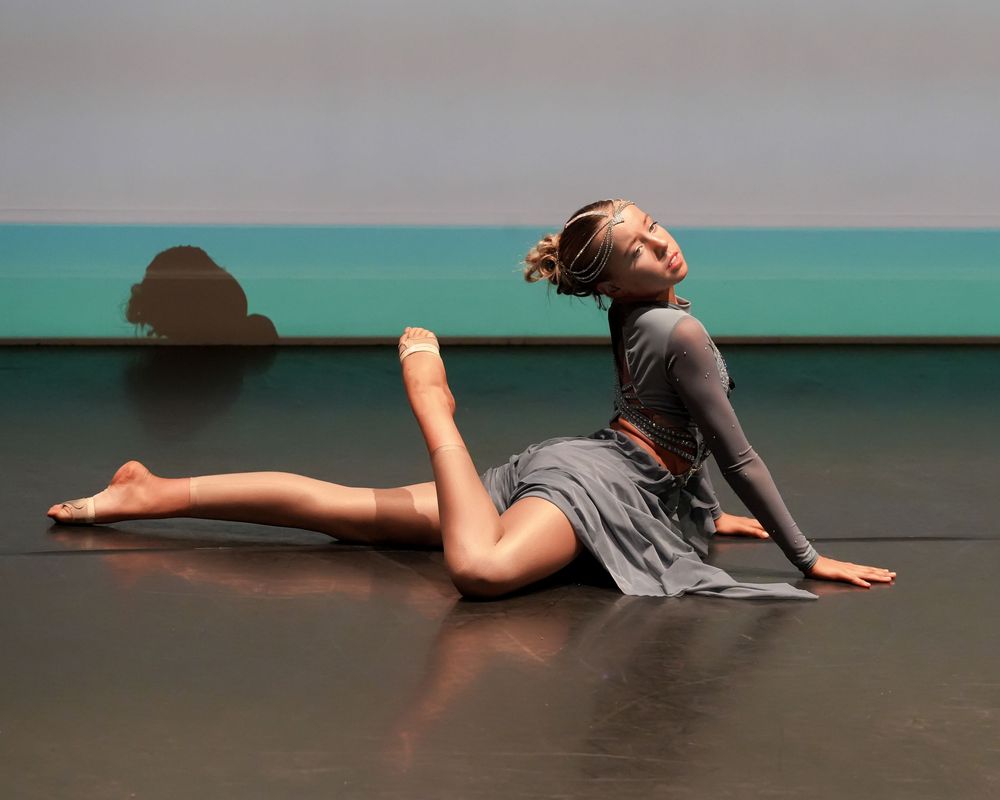 Dancer in gray dress performs expressive floor movement on stage with dramatic back arch pose.