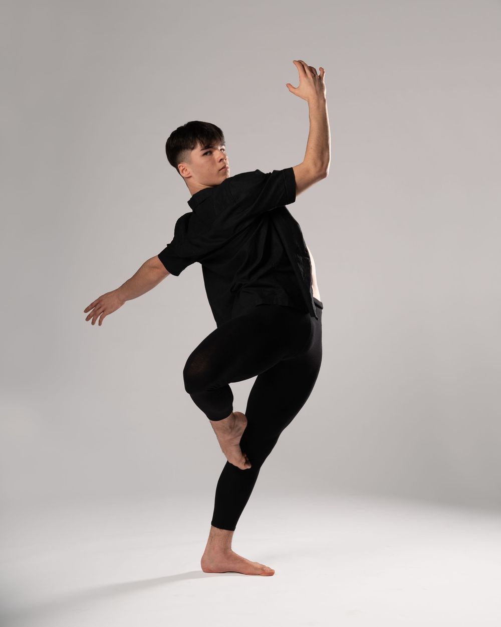 Male dancer in black outfit striking a dynamic pose against a gray background, one knee raised and arm extended.