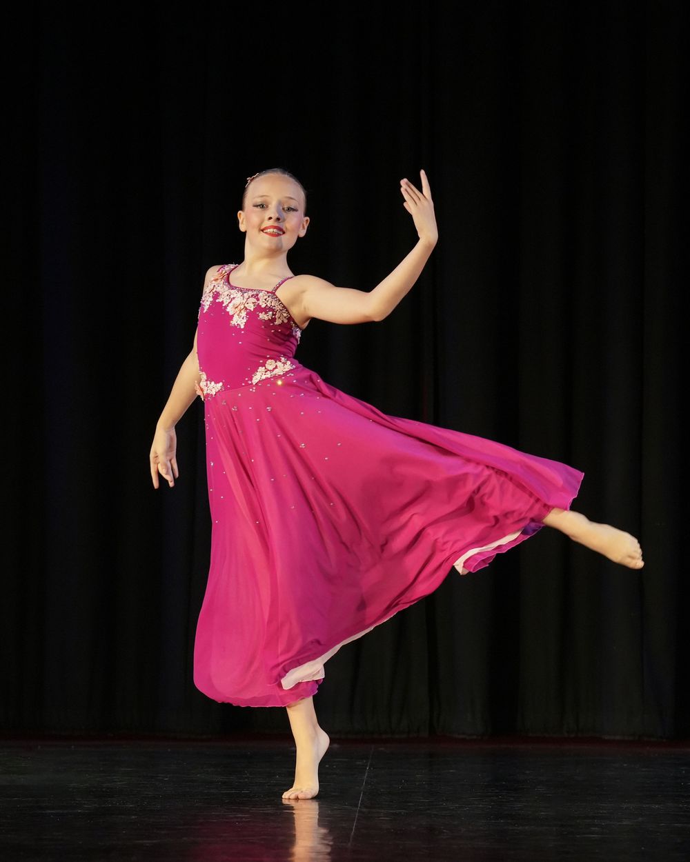 Young girl performs ballet on stage in a flowing magenta gown with floral embroidery under dramatic lighting.