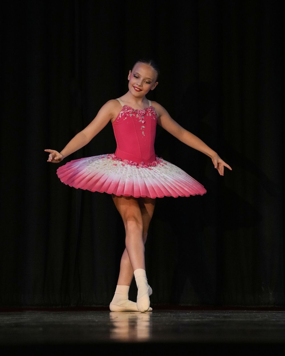 Young ballerina performs on stage in a pink gradient tutu with floral bodice details under bright stage lighting.