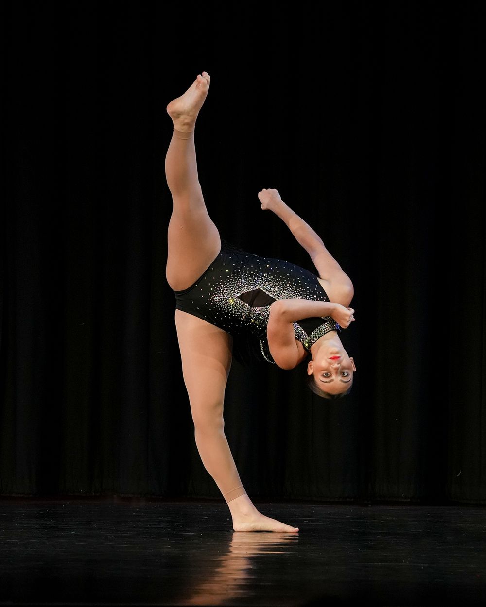 Young gymnast in black sparkly leotard performs a stunning backbend with leg raised high on a dark stage.