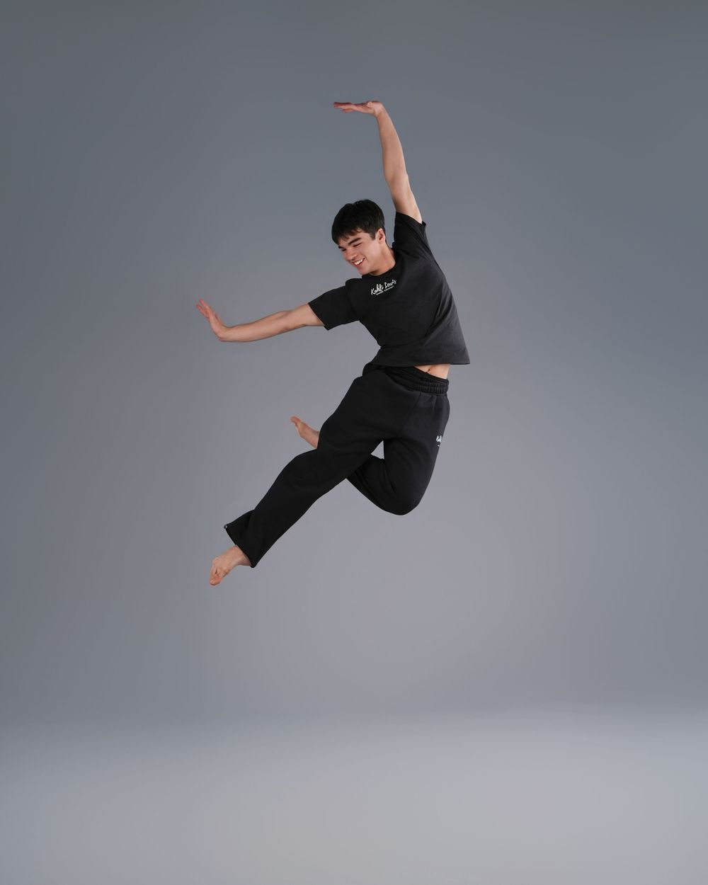 Male dancer in black outfit leaping gracefully mid-air against gray studio background.