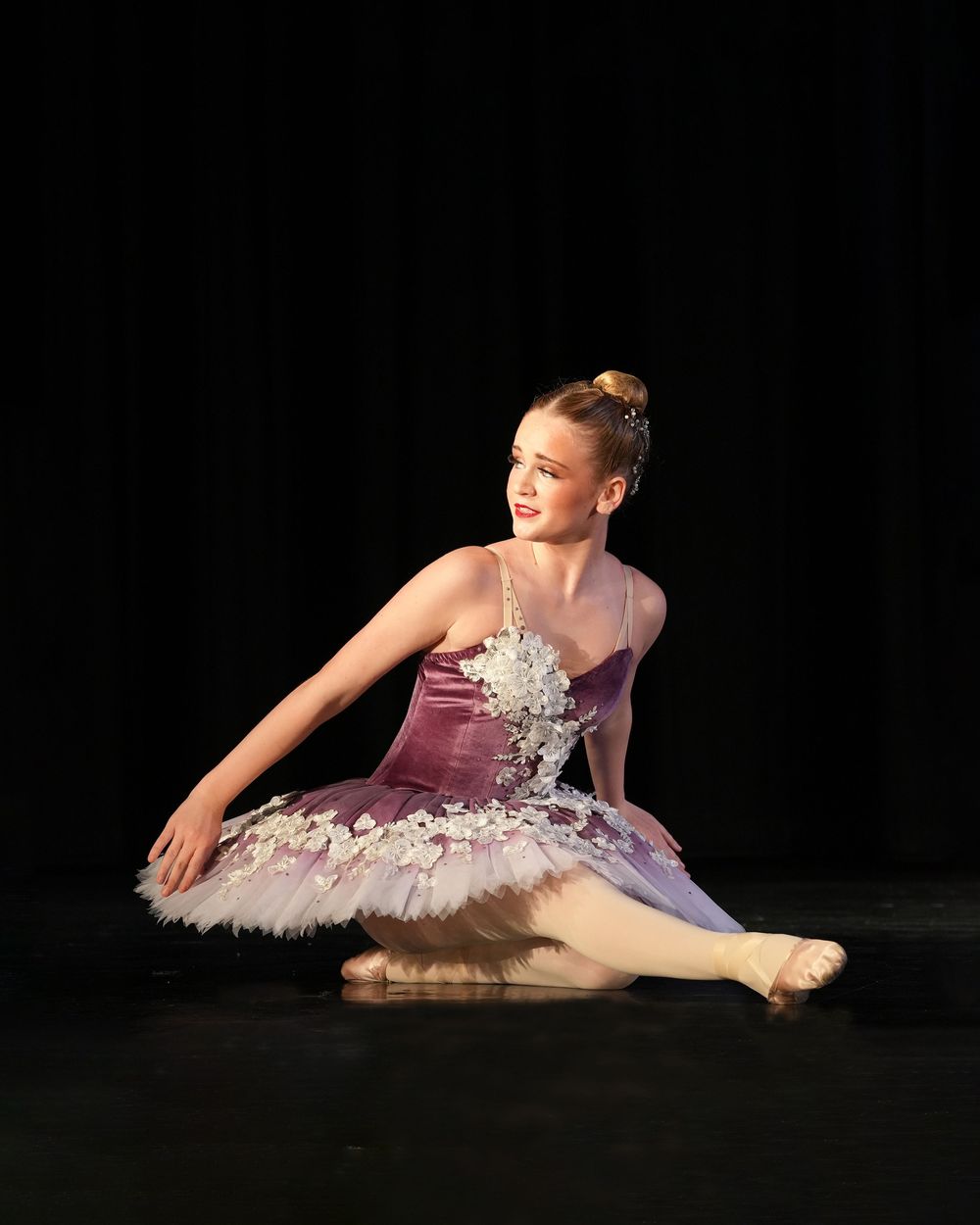 Young ballerina in purple tutu performs graceful floor pose on stage under spotlight during ballet recital.
