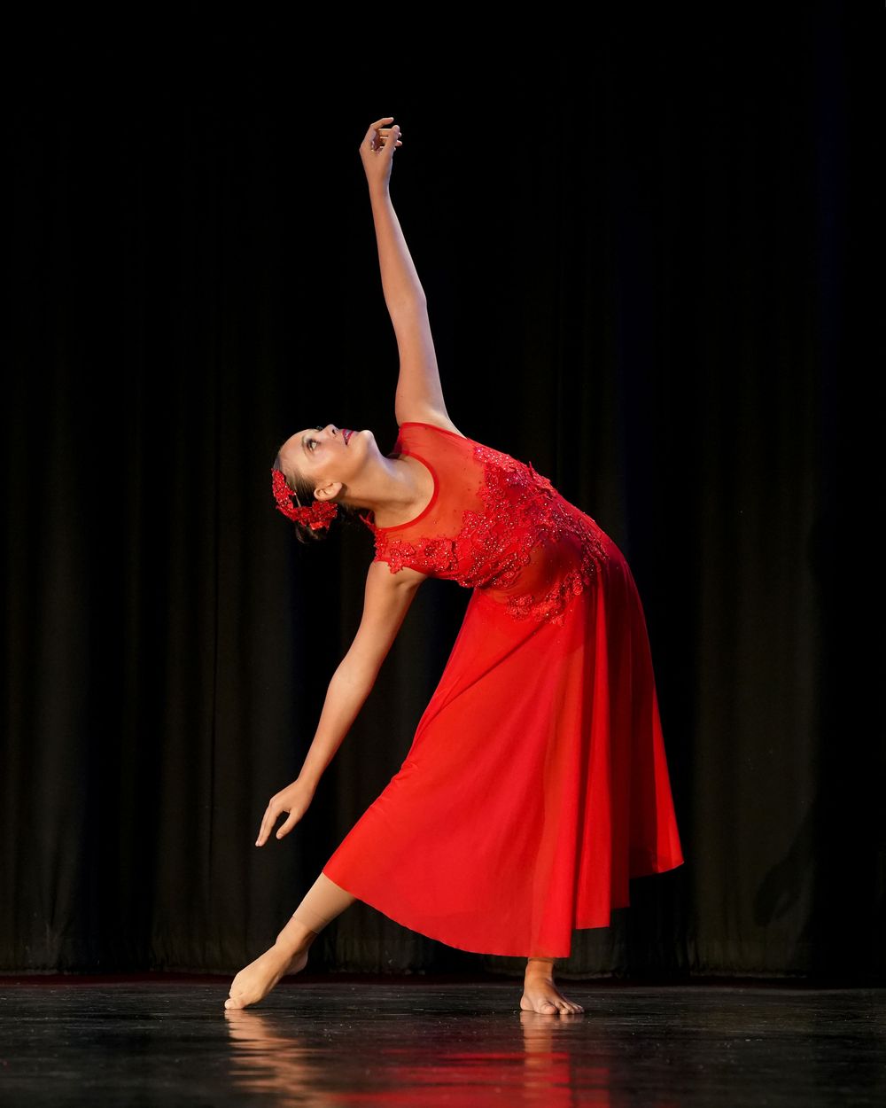 A graceful dancer in a red dress performs an elegant arabesque pose on stage under dramatic lighting.