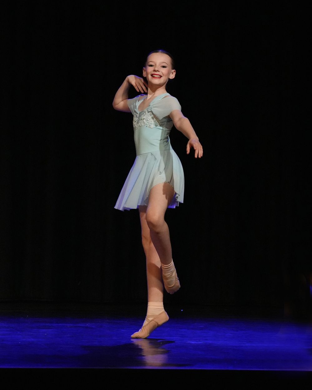 Young girl in light blue ballet costume performs graceful dance routine on stage under spotlight.