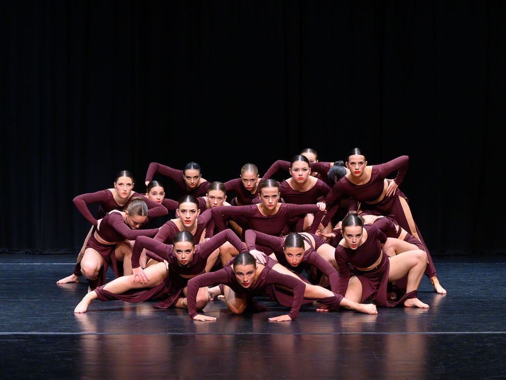 Modern dance ensemble performs synchronized choreography in burgundy costumes on dark stage.