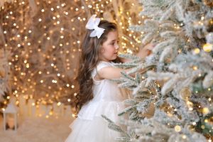 Elegant white dress against snowy Christmas tree with twinkling golden lights in background.