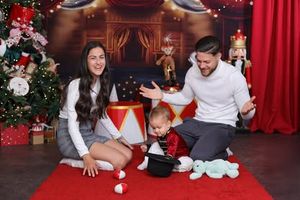 Family playing with toys during festive Christmas photoshoot with red backdrop and decorated tree.