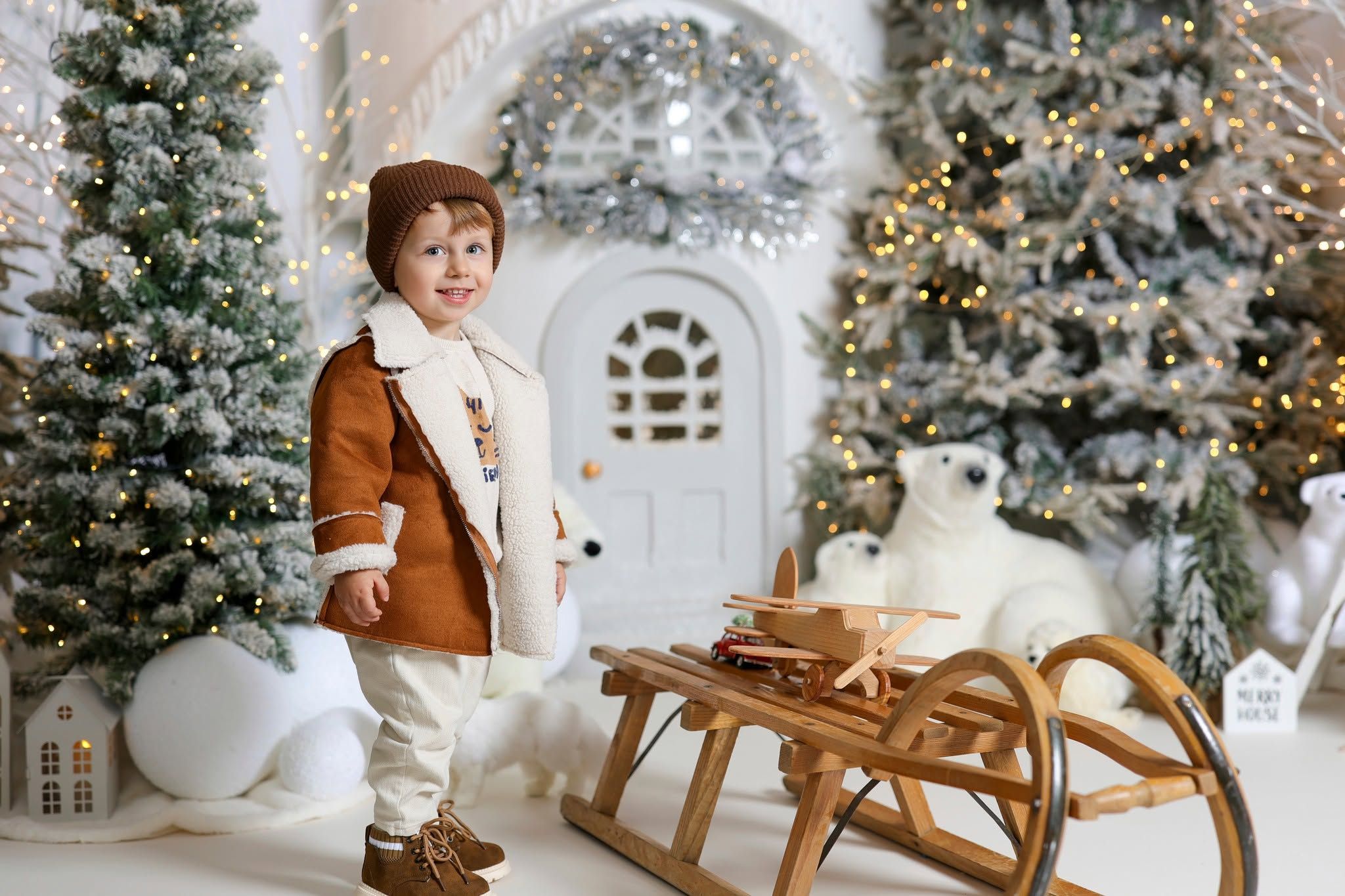 Young child in brown coat stands by wooden sled with polar bear toy amid decorated Christmas trees and white door backdrop.