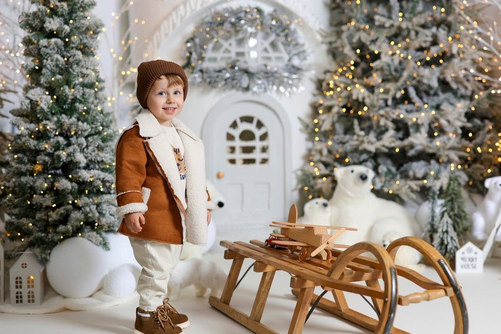 Young child in brown coat stands by wooden sled with polar bear toy amid decorated Christmas trees and white door backdrop.