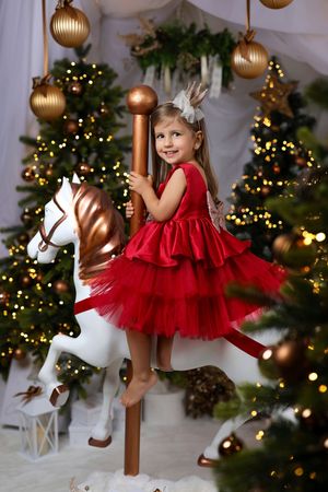 Young girl in red dress rides carousel horse amid Christmas trees with golden ornaments and twinkling lights.