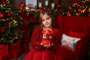 Young girl in red dress holds Christmas decoration near festive tree with bokeh lights.