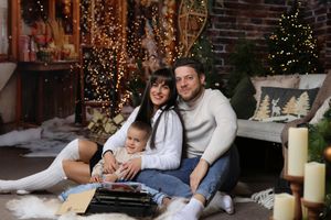 Family of three poses together in festive Christmas decorated room with twinkling lights and holiday decor.