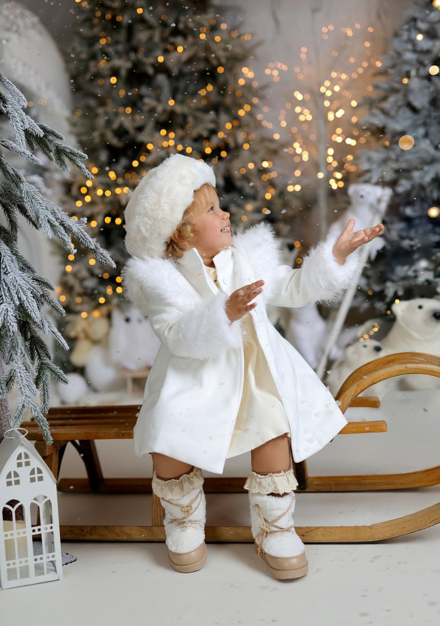 A little girl in white winter coat, fur hat, and boots plays in a festive snowy Christmas scene with bokeh lights.
