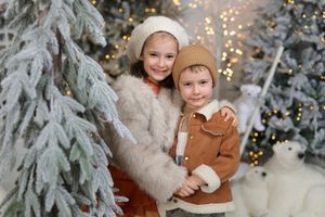 Two children pose together near snow-flocked Christmas trees with warm bokeh lights in a festive holiday photo.