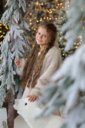 A smiling girl with long hair in a beige fur coat poses with a white bear figurine among snow-covered Christmas trees.