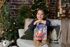 Young child holding a colorful Christmas book, sitting on a sofa near a decorated Christmas tree with warm lights.