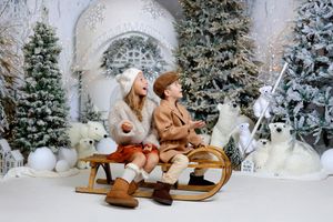 Two children sit on a wooden sled surrounded by snow-covered Christmas trees and polar bear decorations.