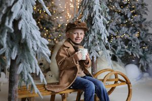 A cheerful boy in a brown coat sits on a wooden sled holding a cup near snowy Christmas trees with warm bokeh lights.