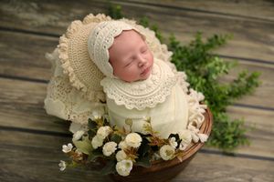 Sleeping newborn in cream lace bonnet and wrap rests peacefully on rustic wood surface with white roses.