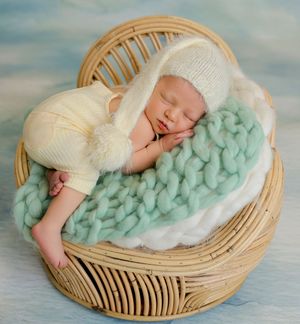 Sleeping newborn baby in cream knit outfit and pompom hat, nestled in a wicker basket with mint chunky knit blanket.