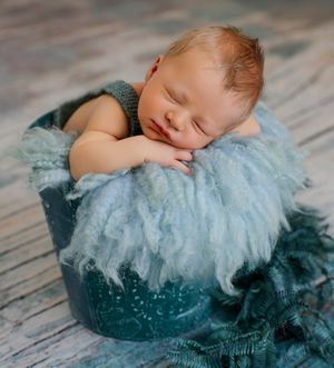Sleeping newborn baby in a teal bucket with fluffy blue wool, wearing a knit romper, perfect newborn photography pose.