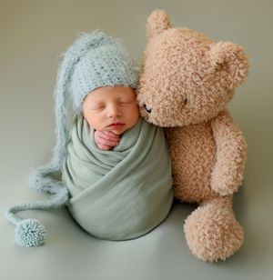Newborn baby swaddled in mint green wrap wearing a blue knit hat, sleeping beside a soft tan teddy bear.