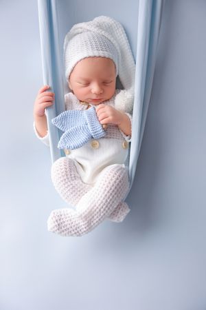 Sleeping newborn in white knit outfit resting in blue hanging prop.