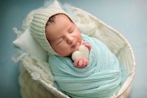 Sleeping newborn wrapped in soft mint green swaddle and knit hat resting in white lace basket.