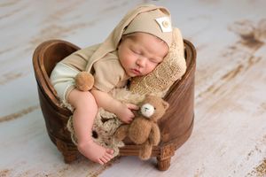 Sleeping newborn baby in beige outfit and hat, cuddling a teddy bear inside a rustic wooden heart-shaped bowl.