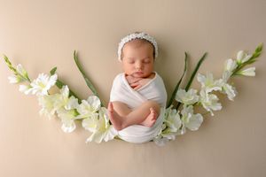 Sleeping newborn baby swaddled in white wrap with floral headband, surrounded by white flowers on beige background.