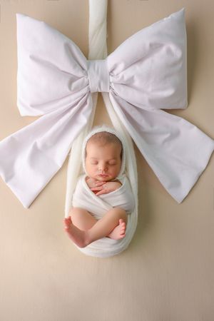 Newborn baby swaddled in white wrap posed as a gift with a large white bow on beige background.