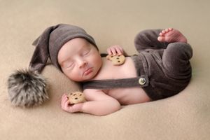 Sleeping newborn in brown outfit with furry pom-pom accessory.