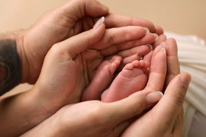 Two adult hands cradling tiny newborn baby feet, with a tattoo visible on one wrist, in a warm close-up photo.