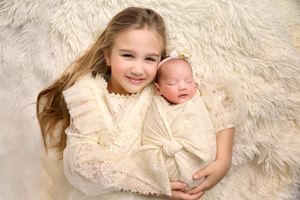 Two siblings in cream lace outfits posed together on fluffy white backdrop.