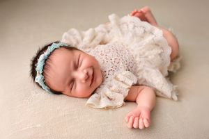 Sweet newborn baby sleeping peacefully in a cream lace dress against a beige background.