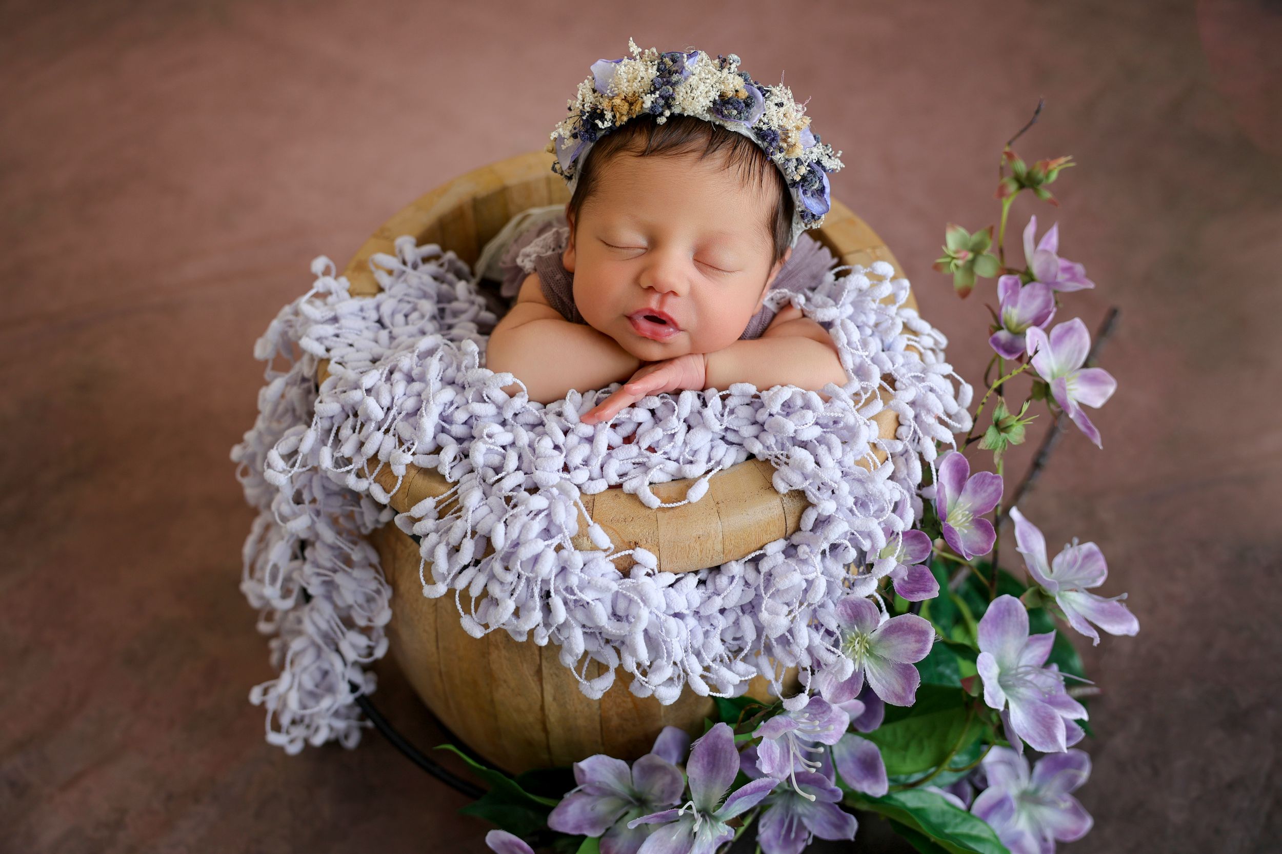 Sleeping newborn baby in wooden bowl with floral bonnet, surrounded by purple flowers and white lace blanket.