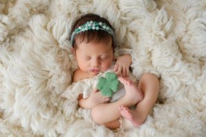 Sleeping newborn baby girl with teal floral headband holding a green four-leaf clover on a white fluffy blanket.